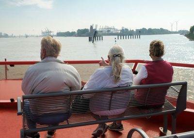 drei ältere Personen sitzen auf dem offenen Oberdeck einer Barkasse und blicken auf den Hafen von Hamburg