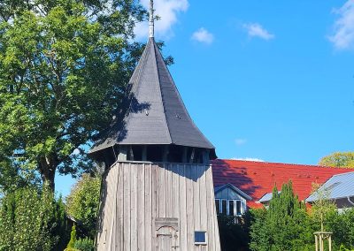 hölzerner Kirchturm der Laurentiuskirche in Langenhorn, Nordfriesland unter strahlend blauem Himmel, im Vordergrund Grabsteine des Friedhofs