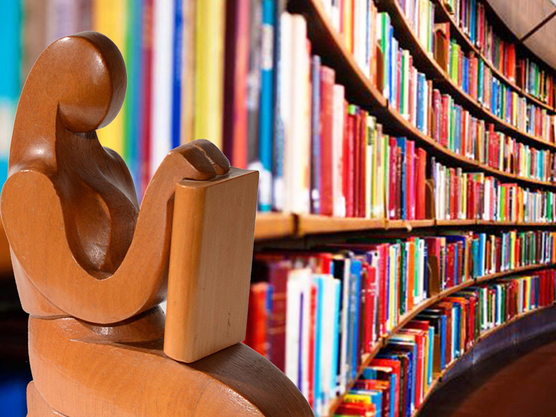 lesende Holzfigur, im Hintergrund Buchregale in einer Bibliothek